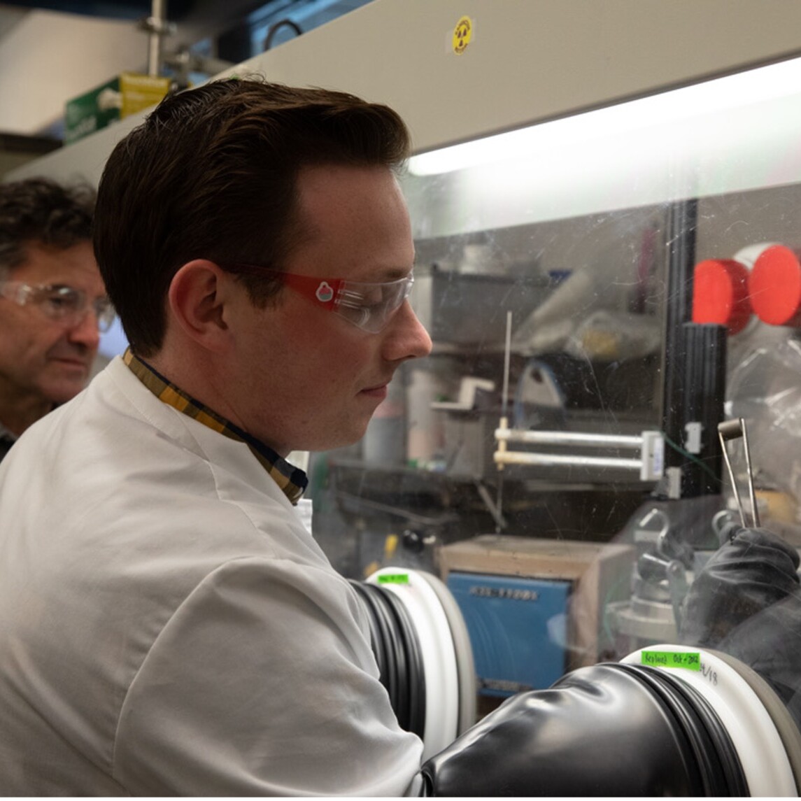 White man with lab coat and lab glasses reaching into glass display