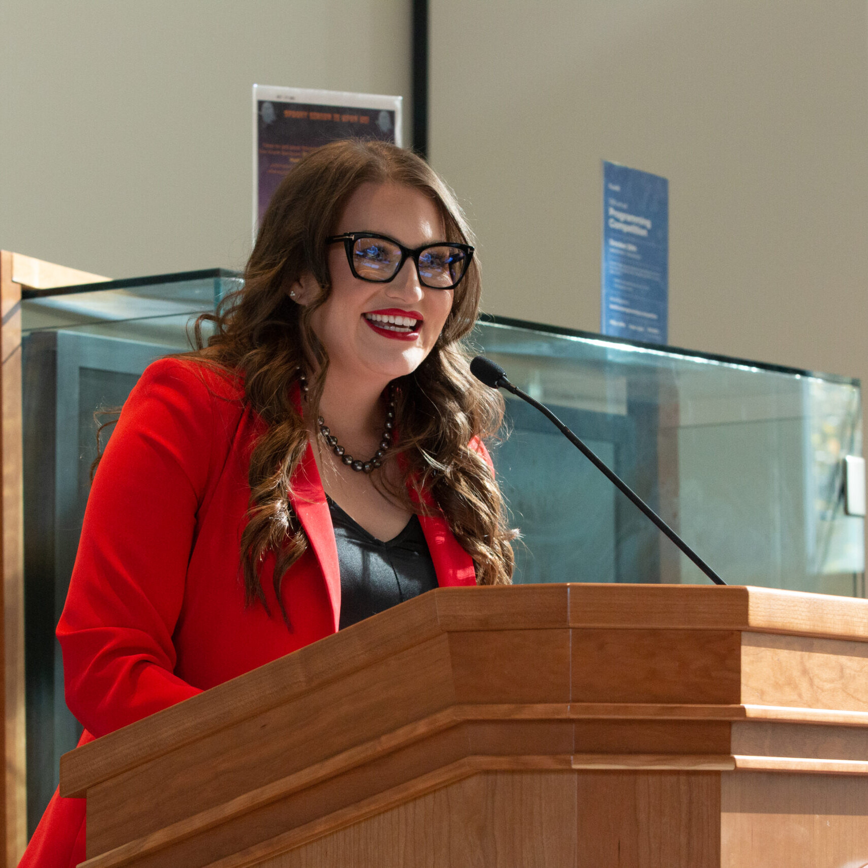 White woman with black glasses wearing a red suit jacket and standing at a podium